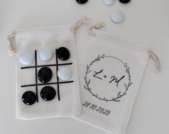 May include: Two off-white drawstring canvas bags. One bag features a black printed tic-tac-toe grid with black and white glass stones placed on it. The other bag displays a black printed wreath design with the letters L + M and the date 28.10.2023.