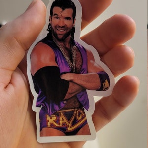 Może przedstawiać: Naklejka z wrestlerem w fioletowym i złotym stroju do zapas&oacute;w. Wrestler ma wąsy i brodę i uśmiecha się. Na naklejce widnieje imię "Razor".