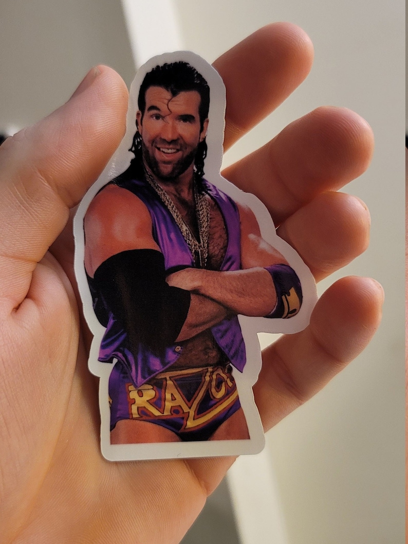 Może przedstawiać: Naklejka z wrestlerem w fioletowym i złotym stroju do zapas&oacute;w. Wrestler ma wąsy i brodę i uśmiecha się. Na naklejce widnieje imię "Razor".