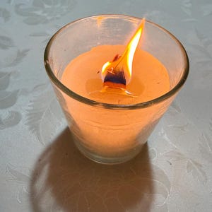 Puede incluir: Una vela naranja encendida en un portavelas de vidrio transparente. La llama es de color amarillo brillante y naranja, proyectando una sombra sobre el mantel blanco. La vela es redonda y tiene una superficie lisa.