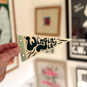 Könnte beinhalten: Ein cremefarbenes Wimpel mit dem Schriftzug "Wingin' it" in Dunkelgrün. Der Wimpel hat einen hellgrünen Rand und Metallösen. Der Wimpel wird von einer Hand mit einem goldenen Ring gehalten.