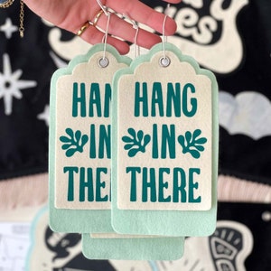 K&ouml;nnte beinhalten: Zwei hellgr&uuml;ne Geschenkanh&auml;nger aus Filz mit wei&szlig;em Hintergrund. Die Anh&auml;nger haben ein blaugr&uuml;nes Blumenmuster und den Text "Hang in there" darauf gedruckt.