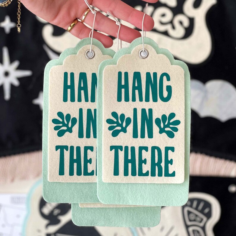 K&ouml;nnte beinhalten: Zwei hellgr&uuml;ne Geschenkanh&auml;nger aus Filz mit wei&szlig;em Hintergrund. Die Anh&auml;nger haben ein blaugr&uuml;nes Blumenmuster und den Text "Hang in there" darauf gedruckt.