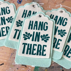 K&ouml;nnte beinhalten: Sechs gr&uuml;ne und wei&szlig;e Filz-H&auml;ngeetiketten mit dem Text "Hang in there" in dunkelgr&uuml;n bedruckt. Die Etiketten sind mit wei&szlig;em Garn gebunden.