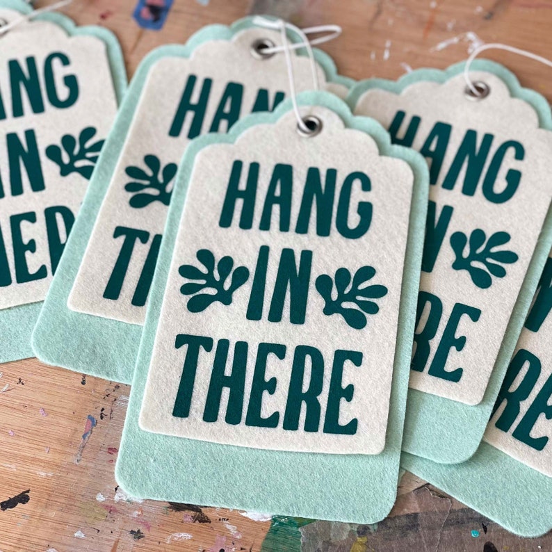 K&ouml;nnte beinhalten: Sechs gr&uuml;ne und wei&szlig;e Filz-H&auml;ngeetiketten mit dem Text "Hang in there" in dunkelgr&uuml;n bedruckt. Die Etiketten sind mit wei&szlig;em Garn gebunden.