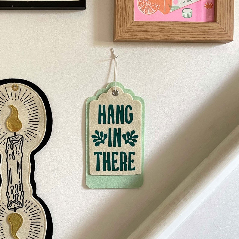 K&ouml;nnte beinhalten: Ein gr&uuml;nes und cremefarbenes Filzetikett mit dem Text "Hang in there" in dunkelgr&uuml;nen Buchstaben. Das Etikett h&auml;ngt an einer Schnur.