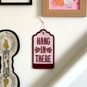 K&ouml;nnte beinhalten: Ein burgunderfarbener Filzanh&auml;nger mit einem rosa Filzeinsatz. Der Anh&auml;nger hat einen gewellten Rand und ein Loch oben. Der Text "HANG IN THERE" ist in Wei&szlig; auf den rosa Filz gedruckt.