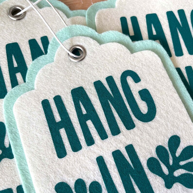K&ouml;nnte beinhalten: Ein Satz wei&szlig;er Filz-Geschenk-Etiketten mit einem gewellten Rand und einer gr&uuml;nen Aufschrift "HANG IN". Jede Etikette hat oben ein Loch und eine silberne Metall&ouml;se.