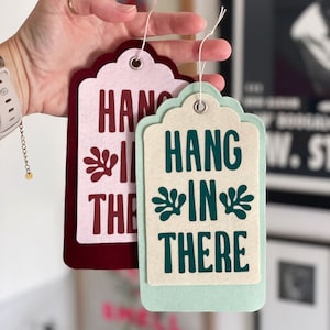 K&ouml;nnte beinhalten: Zwei Filz-H&auml;ngeetiketten mit dem Text "Hang in there" in verschiedenen Farben und Schriftarten. Das Etikett links ist burgunderrot mit rosa Beschriftung und einem gr&uuml;nen Blattdesign. Das Etikett rechts ist hellgr&uuml;n mit wei&szlig;er Beschriftung und einem gr&uuml;nen Blattdesign.