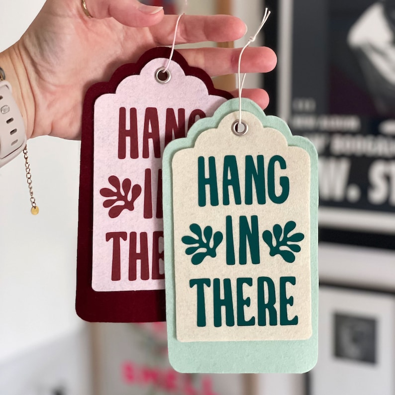 K&ouml;nnte beinhalten: Zwei Filz-H&auml;ngeetiketten mit dem Text "Hang in there" in verschiedenen Farben und Schriftarten. Das Etikett links ist burgunderrot mit rosa Beschriftung und einem gr&uuml;nen Blattdesign. Das Etikett rechts ist hellgr&uuml;n mit wei&szlig;er Beschriftung und einem gr&uuml;nen Blattdesign.
