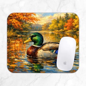 Op de afbeelding: Een rechthoekige muismat met een wilde eend die op een kalm meer zwemt. De scène toont herfstbladeren in oranje, gele en groene tinten die reflecteren op het wateroppervlak. Een witte computermuis ligt aan de rechterkant van de muismat.