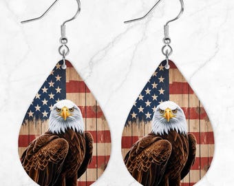 May include: Teardrop earrings with a bald eagle design. The background features the American flag on a wooden texture. The eagle has brown feathers, a white head, and a yellow beak. The earrings have silver-coloured hooks.