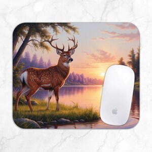 Puede incluir: Alfombrilla de ratón rectangular con un paisaje sereno. Un ciervo con grandes cuernos se encuentra junto a un lago al atardecer. Un ratón de ordenador blanco está al lado. La escena transmite tranquilidad.