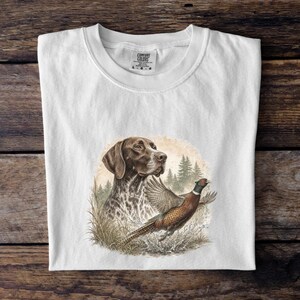 Puede incluir: Una camiseta blanca con un estampado gráfico de un perro de muestra alemán de pelo corto marrón y blanco y un faisán en vuelo. El diseño incluye un fondo de bosque con pinos y hierba alta. La camiseta tiene una etiqueta de talla L en el cuello.