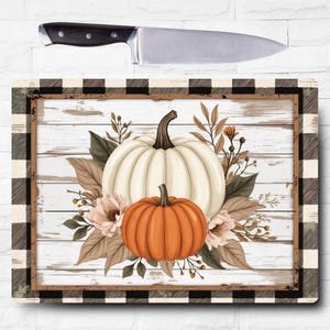 May include: A decorative cutting board featuring a white and orange pumpkin design with floral accents. The board has a black and white buffalo check border and a stainless steel chef's knife.
