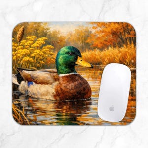 Op de afbeelding: Een rechthoekige muismat met een gedrukte illustratie van een wilde eend met een groene kop, gele snavel en bruine borst die in het water zwemt. De achtergrond toont een levendig herfstlandschap met goudgeel gebladerte en hoog gras.