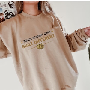 May include: A tan sweatshirt with the text "POLICE ACADEMY GRAD BUILT DIFFERENT" in gold lettering. The design includes a star and badge graphic. The sweatshirt is a neutral color and has long sleeves.