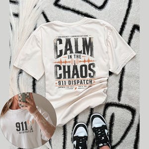 Pode incluir: Camiseta creme com o texto "CALM IN THE CHAOS" e "911 DISPATCH" em uma fonte desgastada. A camiseta também apresenta um gráfico de onda sonora e uma torre de rádio. Tênis preto e branco são visíveis.