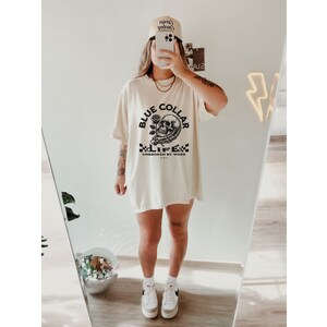 May include: A white oversized t-shirt with a black skull and rose graphic. The graphic says "Blue Collar Life" and "Unbroken by Work". The model is wearing a white baseball cap with the text "Cowboy Country" on it.