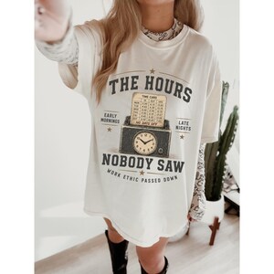 Può includere: Una t-shirt color bianco sporco con la grafica di un orologio marcatempo e un cartellino segnatempo con il testo THE HOURS NOBODY SAW, EARLY MORNINGS, LATE NIGHTS, NO DAYS OFF e WORK ETHIC PASSED DOWN. Il design include stelle e un orologio vintage.