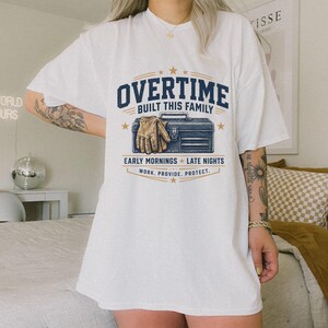 Può includere: Una t-shirt bianca a maniche corte con una grafica raffigurante una cassetta degli attrezzi blu e un guanto da lavoro beige. Il testo recita: OVERTIME BUILT THIS FAMILY, EARLY MORNINGS, LATE NIGHTS, WORK. PROVIDE. PROTECT. Il design include stelle dorate.