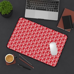 May include: A red mousepad with a repeating white pattern of Greek letters, including Delta, Sigma, and Theta. A white computer mouse sits on the pad, with a laptop, notebook, and coffee cup in the background.