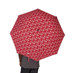 May include: Red umbrella with a repeating white pattern of Greek letters. The umbrella is open, displaying its full canopy. The handle is visible, and the background is white.