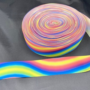 May include: A roll of rainbow ombre ribbon with a blue, yellow, red, and orange color gradient. The ribbon is rolled up and has a white backing.