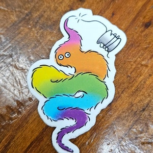 May include: A rainbow-coloured fuzzy worm with a small, grey, cylindrical object on its head. The worm is curled up in a circle.