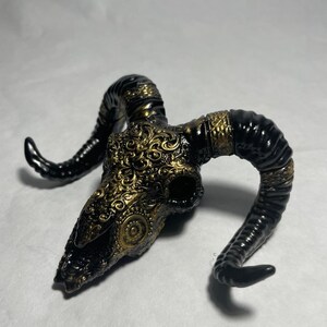 May include: A black and gold ram skull with intricate gold detailing. The skull is a decorative object with a rustic and gothic aesthetic.