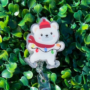 May include: A white polar bear wearing a red Santa hat and a red scarf with colorful Christmas lights. The bear is a charm on a clear plastic retractable badge holder.