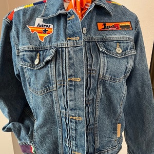 Peut inclure: Veste en jean bleue avec des écussons, dont un écusson "Astros" en forme du Texas et un écusson de train. La veste a des bordures colorées sur les manches et les épaules, et un bandana orange est drapé sur le col.