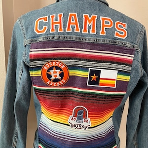May include: A denim jacket with the word "CHAMPS" in orange letters across the back. The jacket features a colorful striped fabric panel and patches with the Houston Astros logo, Texas flag, and a "2022 Astros" patch.