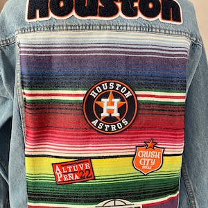 Peut inclure: Veste en jean bleu clair avec un panneau arrière rayé coloré. Les écussons incluent "Houston" sur les épaules, un logo des Astros de Houston, "Crush City Texas", "Altuve Pena 22" et un graphique de stade. Veste unique et élégante.