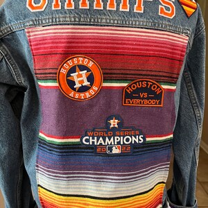 May include: A denim jacket with a colorful woven back panel. The jacket features patches with the text "CHAMPS", "Houston Astros", "Houston vs Everybody", and "World Series Champions 2022". The cuffs have colorful fabric accents.