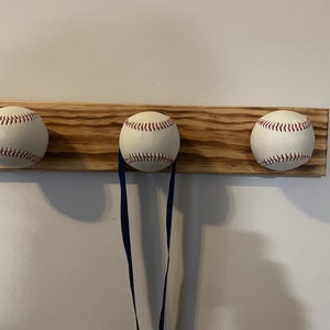 Op de afbeelding: Een houten wandbevestigde kapstok met drie baseballs als haken. De kapstok is lichtbruin gebeitst en heeft een natuurlijke houtnerf.