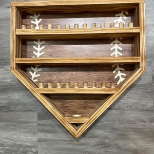May include: Wooden baseball home plate display shelf with three shelves and small wooden pegs. The interior features baseball stitching designs. The frame is made of light brown wood, and the interior is a darker brown.