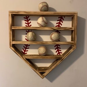 May include: A wooden baseball-shaped shelf with three shelves to display baseballs. The shelf is painted white with red stitching details. Five baseballs are displayed on the shelves.