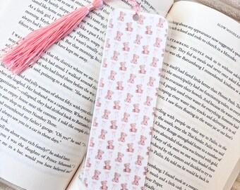 Puede incluir: Un marcapáginas con un patrón de pequeños ositos de peluche rosas y lazos rosas. Tiene una borla rosa y está sobre un libro abierto.