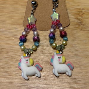 Può includere: Un paio di orecchini stravaganti con ciondoli a forma di unicorno bianco con criniere e code rosa, gialle e blu. Gli orecchini hanno fili di perline in vari colori, tra cui perline rosa, viola e iridescenti, e sono sormontati da accenti a forma di stella.