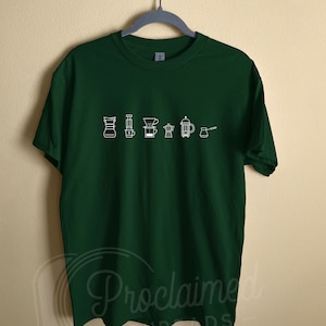 Peut inclure: Un t-shirt vert foncé avec un motif blanc représentant différentes méthodes de préparation du café, notamment un filtre à café, une cafetière à piston et une cafetière italienne.