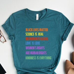 May include: Teal t-shirt with the text "BLACK LIVES MATTER, SCIENCE IS REAL, NO HUMAN IS GLOBAL, LOVE IS LOVE, WOMEN'S RIGHTS ARE HUMAN RIGHTS, KINDNESS IS EVERYTHING" in rainbow colors. The shirt is styled with accessories and shoes.