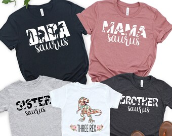 May include: A set of four t-shirts featuring a dinosaur theme. The shirts are in black, grey, white, and dark grey. The shirts have the text "Dada saurus", "Mama saurus", "Sister saurus", and "Brother saurus" printed on them. One shirt features a floral dinosaur with the text "Three Rex".