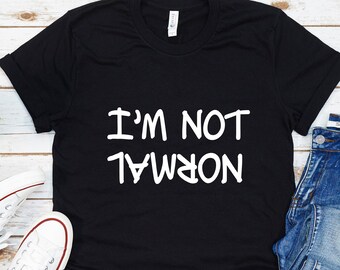 May include: Black t-shirt with white text that reads "I'M NOT NORMAL" in a playful, upside-down font. The shirt is displayed on a white wood background with a pair of white sneakers and blue jeans.