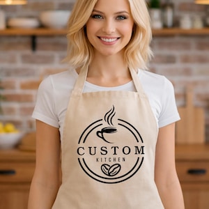 May include: A beige apron with a black circular logo that says "CUSTOM KITCHEN" with a coffee cup and coffee bean design. The apron has two front pockets and is being worn in a kitchen setting.