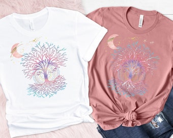 May include: Two t-shirts with a colorful tree of life design. The white t-shirt has a pink moon and gold glitter. The pink t-shirt has a gold crescent moon and stars. Both shirts feature a tree of life with roots and branches.