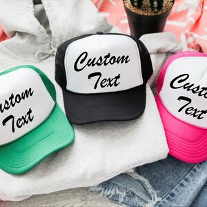 May include: Three trucker hats with white fronts and mesh backs. One hat is green, one is black, and one is pink. Each hat has the words "Custom Text" in black script across the front.