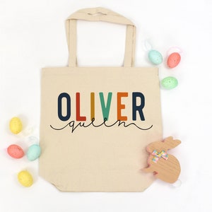 May include: A beige canvas tote bag with the name "OLIVER queen" printed in colorful letters. The bag has two handles and is surrounded by pastel-colored Easter eggs and a wooden bunny decoration.