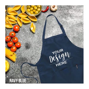 May include: Navy blue apron with the text "YOUR Design HERE" in white script. The apron has a pocket and a loop for hanging. The words "NAVY BLUE" are at the bottom left. The background includes pasta, tomatoes, and olives.