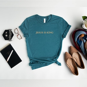 Pu&ograve; includere: Maglietta blu turchese con il testo "JESUS IS KING" in lettere dorate.